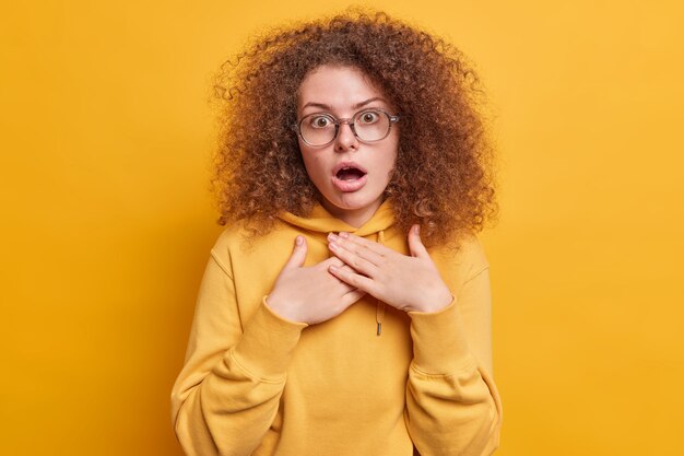 Shocked stupefied curly haired woman hears amazing news keeps hands on chest gasps and stares  wears big optical glasses and sweatshirt isolated on yellow wall. Reaction concept
