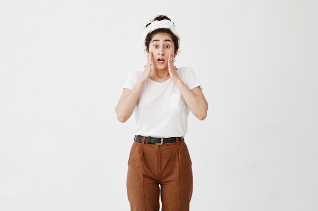 Shocked stupefied young female in white t-shirt and brown trousers looks with popped dark eyes and opened mouth, keeps hands on cheeks, doesn`t believe in her failure, has puzzled frustrated look