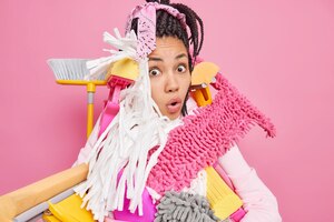 shocked stupefied young woman overloaded with cleaning tools has much work about house ready for tidying up apartment poses against pink background spring cleaning and housekeeping concept