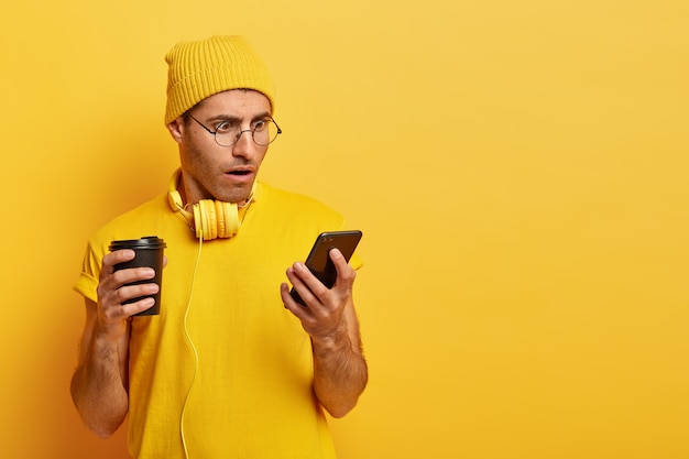Шокирован стильный парень в желтом наряде и очках, использует смартфон