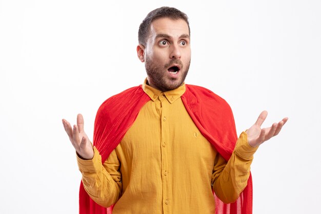 Shocked superhero man with red cloak holds hands open looking at side isolated on white wall