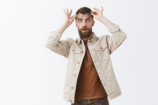 Shocked and surprised bearded man in glasses posing against the white wall