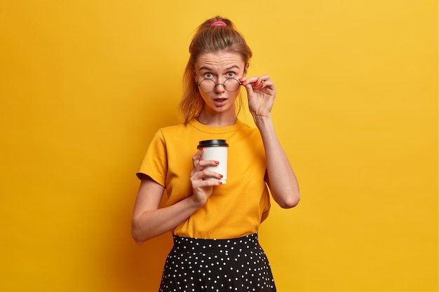 Shocked surprised European female student looks through glasses, poses with takeaway coffee, hears astonishing news, cannot believe her eyes, wears casual clothes