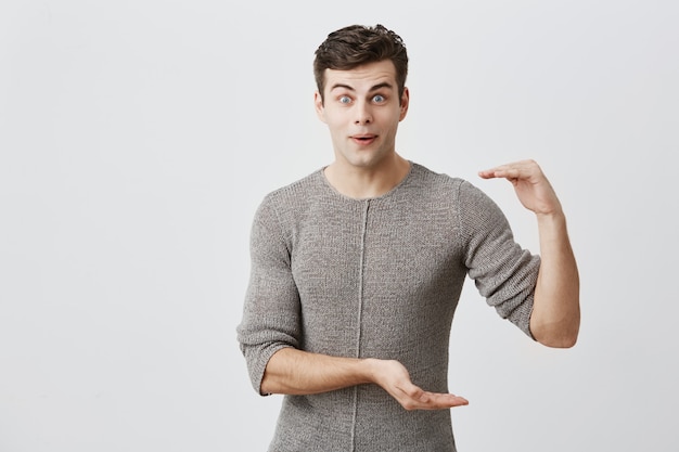 Shocked surprised excited European handsome male model wearing sweater with dark hair and bugged eyes showing size of something with hands, gesturing. Body language concept