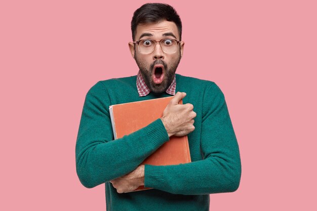 Shocked surprised unshaven young man carries vintage book closely to chest, stares with bugged eyes, has scared expression, wears optical glasses