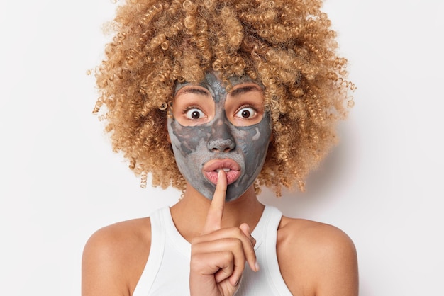 Shocked surprised woman makes hush gesture takes care of dry complexion applies beauty clay mask spreads rumors dressed casually isolated over white background. Face care procedures at morning