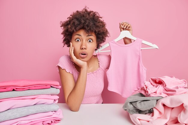 Shocked surprised woman with curly hair holds t shirt on hanger folds laundry at home does household duties cleans out closet sits at table isolated on pink 