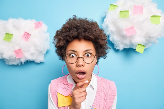 Shocked terrified female student uses important note uses paper stickers post keeps hand on chin being deeply surprised wears round spectacles formal clothes isolated over blue wall