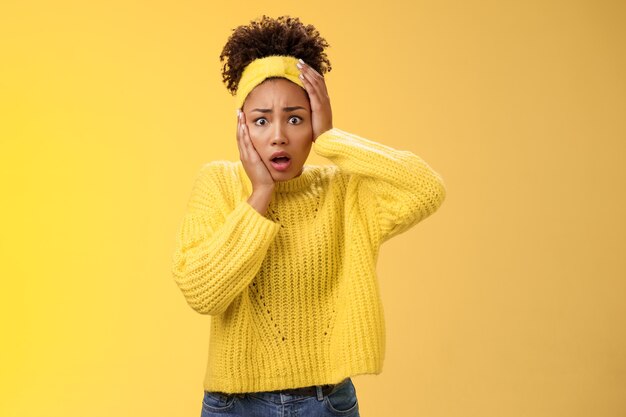 Shocked terrified timid insecure young stylish african-american woman panic touch face concerned gasping open mouth widen eyes frightened bad consequences standing upset nervous yellow background.