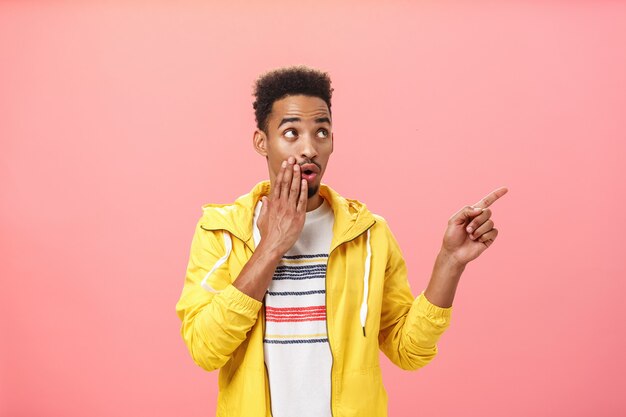 Shocked thrilled african american male with beard and afro haircut saying wow with opened mouth and palm on lip pointing and gazing at upper right corner intrigued over pink background