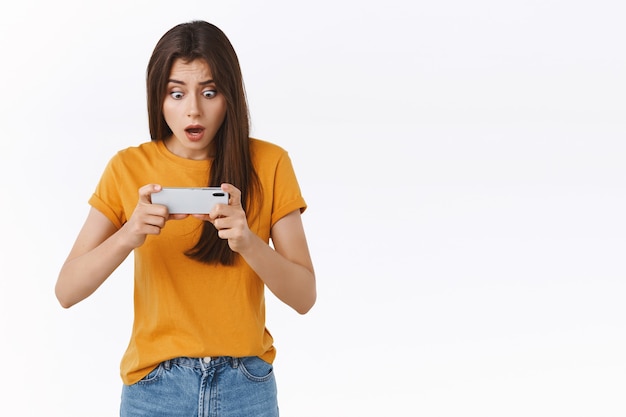 Shocked thrilled young brunette female playing intense battle game, holding smartphone horizontally tap mobile screen to win, gasping stare telephone display with popped eyes, white background