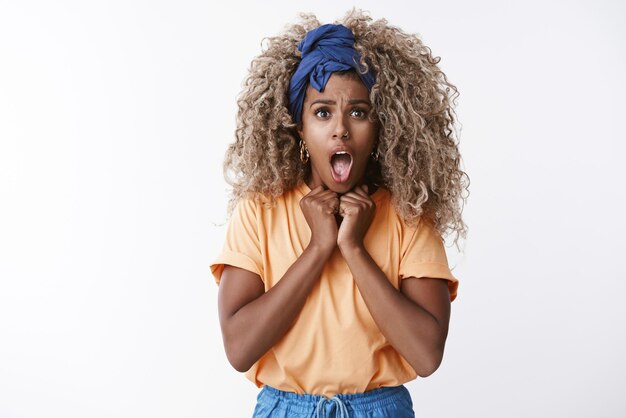 Shocked timid and innocent cute hipster africanamerican female with blond afro hair orange tshirt gasping shocked and scared open mouth press hands to chest intense looking worried