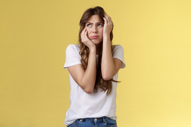 Shocked timid insecure young panicking woman wanna cry standing anxious frightened grab head both hands freak out look away praying for help terrified stand yellow background horrified