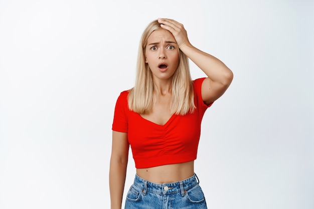 Shocked and troubled blond woman hold hand on head, staring frustrated on white, standing on white.