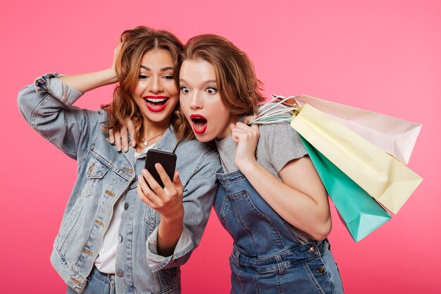 Shocked two women friends holding shopping bags using mobile phone.
