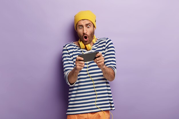 Shocked unshaven guy holds mobile phone horizontally, holds breath, plays games online, being addicted, wears yellow hat and striped jumper