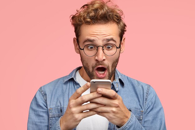 Shocked unshaven man looks surprisingly and with terrified expression at screen of smart phone, uses free wifi for messaging in social networks, reads awful news via internet. Omg concept