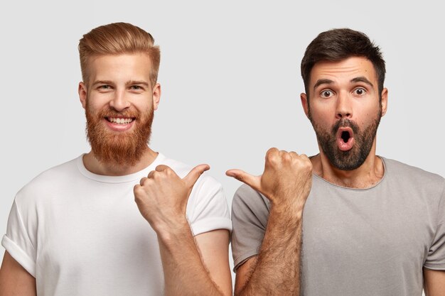 Shocked unshaven man with dark stubble indicates at his companion, wears grey t shirt. Cheerful ginger guy with trendy haircut and bristle points at fellow. Two friends model indoor over white wall