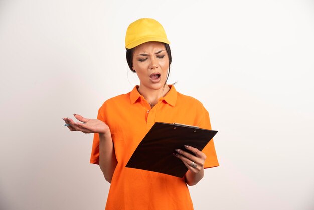 Shocked woman courier with pencil looking on clipboard on white wall.