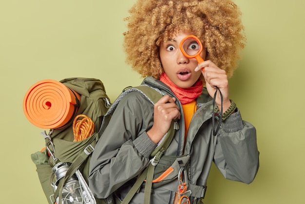 Free photo shocked woman explorer holds magnigying glass over eye cannot believe own eyes explores new place dressed in activewear carries rucksack isolated over green background tourism and camping concept