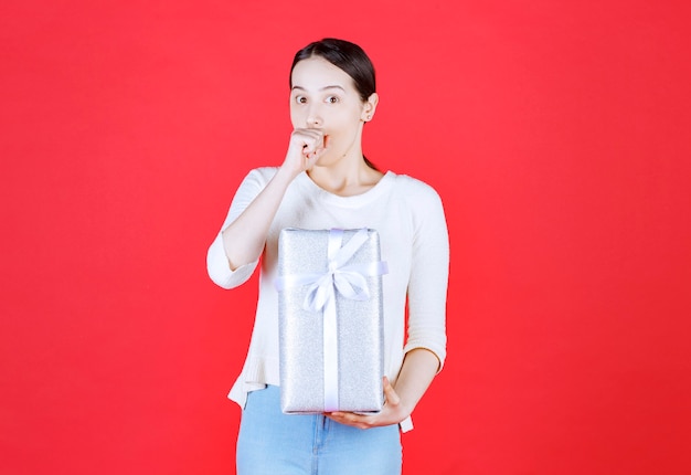 Shocked woman holding gift box on red wall