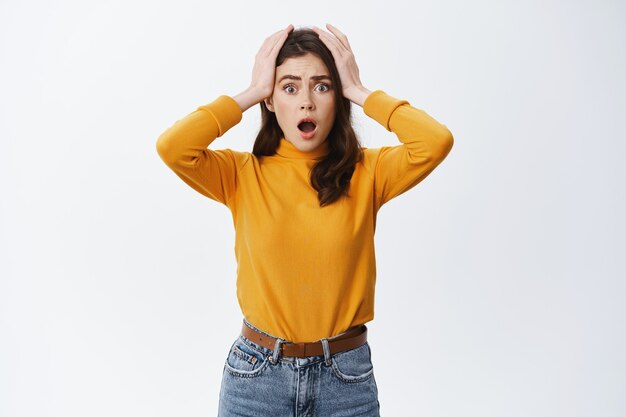 Shocked woman holding hands on head with concerned and anxious face, feeling troubled, facing big problem and looking indecisive at front, white wall