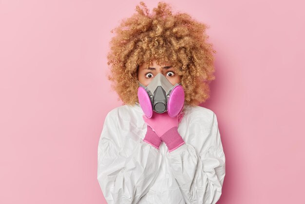 Shocked woman keeps hands on neck suffers from suffocation breathes dangerous gases or chemicals poured on ground wears white safety suit and respirator isolated over pink wall Ecological problem