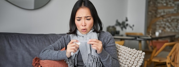 Free photo shocked woman looks concerned at her thermometer catching cold has fever sitting on sick leave at