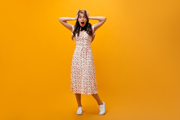 Shocked woman in midi dress looks into camera. Curly girl in white summer outfit and sneakers posing on orange background.