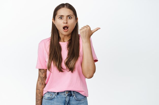 Shocked woman pointing finger right, looking displeased at camera, complaining on advertisement, white background.