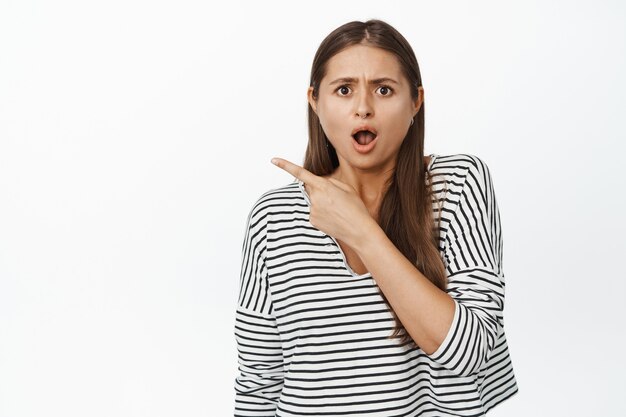 Shocked woman pointing at upper left corner and looking concerned, startled with something aside on white.