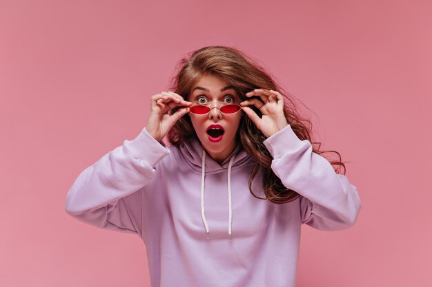 Shocked woman in purple oversized hoodie takes off red sunglasses