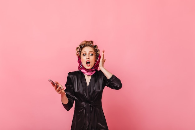 Shocked woman in scarf holding smartphone on pink wall