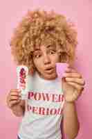 Free photo shocked woman stares at unfamiliar hygiene product holds menstruation cup and sanitary napkin has critical days dressed in casual t shirt isolated over pink background feminine health care