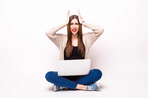 Foto gratuita donna scioccata in t-shirt e occhiali da vista seduta sul pavimento con un computer portatile e guardando la telecamera su grigio