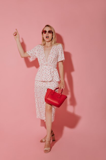 Shocked woman in white clothes red sunglasses and stylish shoes holding red handbag and pointing to place for text on pink backdrop