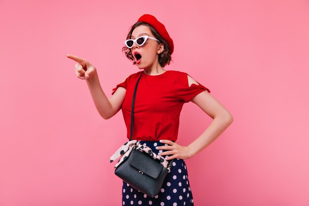 Shocked woman in white glasses posing in red beret. Emotional french lady standing.