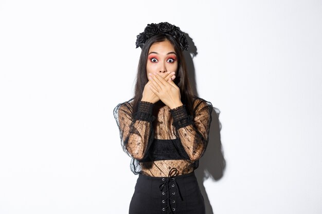 Shocked woman in witch outfit gasping and cover mouth speechless, standing over white background, looking at something surprising, wearing black lace dress and wreath, white background.