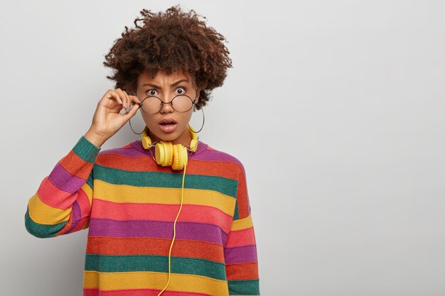 Shocked woman with Afro hairstyle, looks through round spectacles, opens mouth, has stupefied gaze, wears headphones around neck