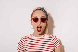 Free photo shocked woman with red lips in sunglasses, cool earrings and striped modern t-shirt looking into camera on isolated wall