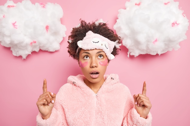 Shocked wondered Afro American woman with pads under eyes points upwards impressed by incredible thing keeps jaw dropped wears nightclothes blindfold on forehead shows something on white clouds