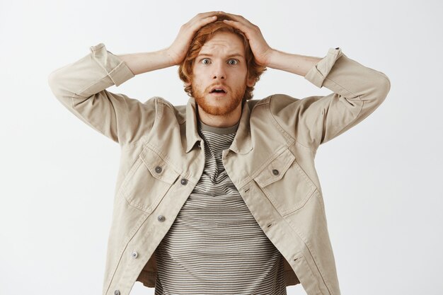 Shocked and worried bearded redhead guy posing against the white wall