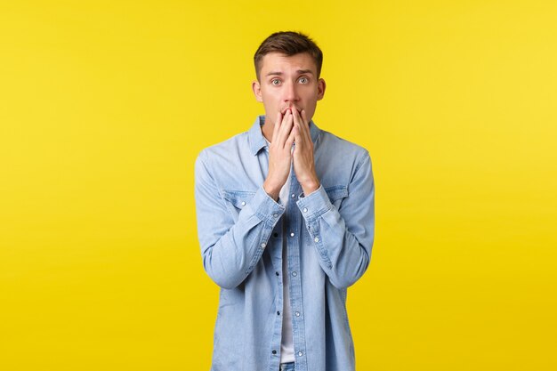Shocked and worried blond man looking concerned at camera, touching lips and stare anxious at camera, hear about terrible accident, express compassion and empathy, yellow background.