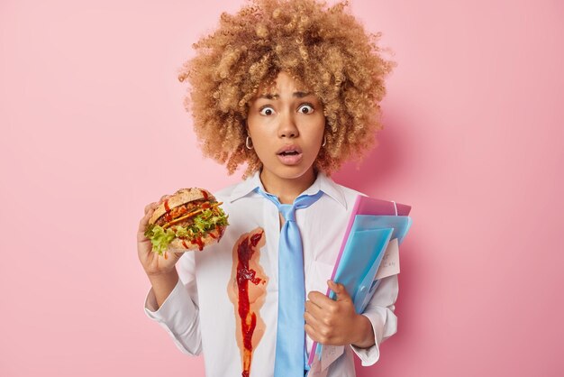 Shocked worried female schoolgirl eats unhealthy food holds hamburger and folders stares at camera wears dirty shirt and tie being regardless while having snack isolated over pink background