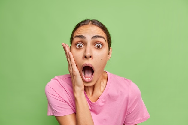 Free photo shocked worried young european woman keeps jaw dropped hand on cheek cannot believe in amazing news wears casual pink t shirt isolated over green wall feels very surprised