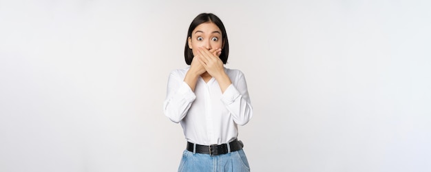 Shocked young asian girl close mouth cover lips with hands and looking amazed standing over white background Taboo gesture