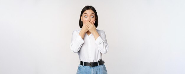 Shocked young asian girl close mouth cover lips with hands and looking amazed standing over white background Taboo gesture