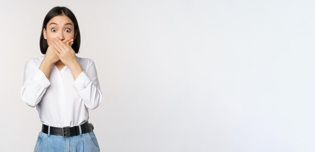 Shocked young asian girl close mouth cover lips with hands and looking amazed standing over white background