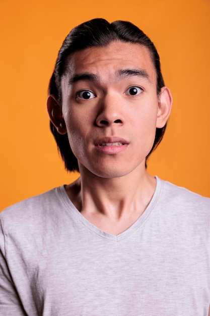 Shocked young asian man facial expression closeup. Surprised teenager with fear emotion on face looking at camera, scared attractive person portrait close view on orange background