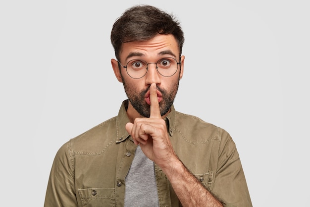 Shocked young bearded male makes silence sign, looks secretly, being frightened by unexpected rumors, isolated over white wall. Shh, be silent! Secrecy and body language concept